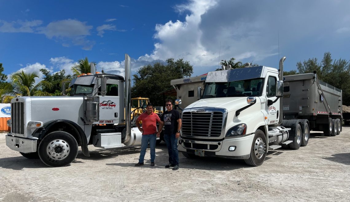Two men talking beside large trucks under a partly cloudy sky.