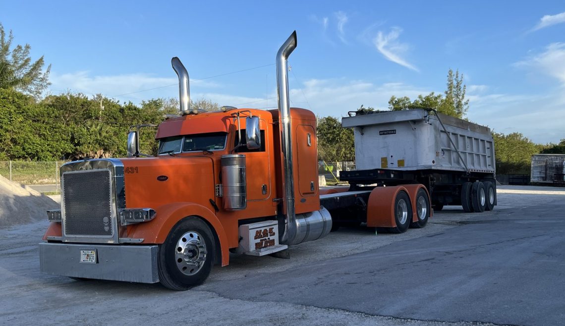 Orange semi-truck with trailer parked on a gravel road under a clear blue sky.